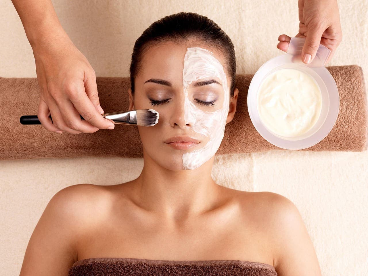 A facialist brushing a cream mask onto a guest's face as she rests on a folded towel