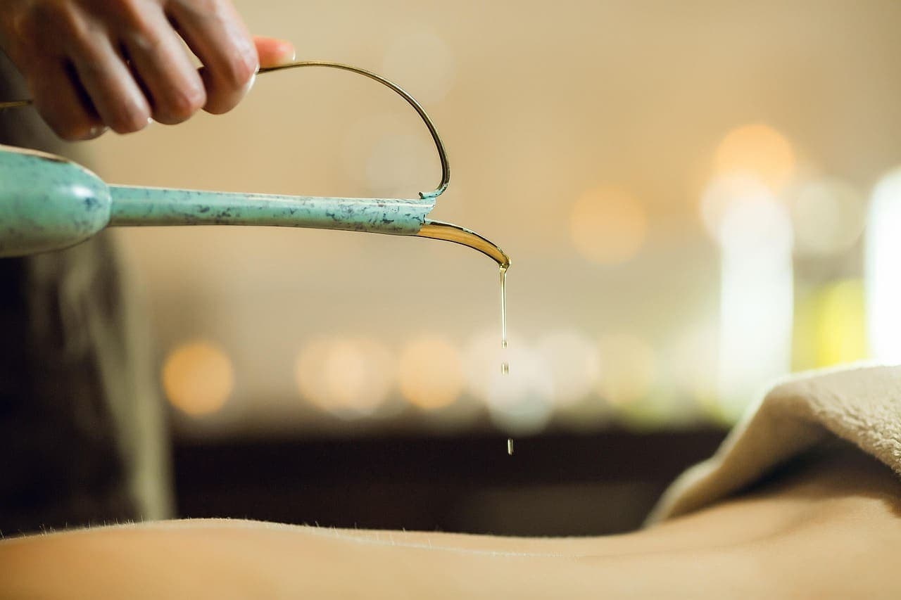 Warm massage oil poured in a thin stream from a copper spout onto a guest's back