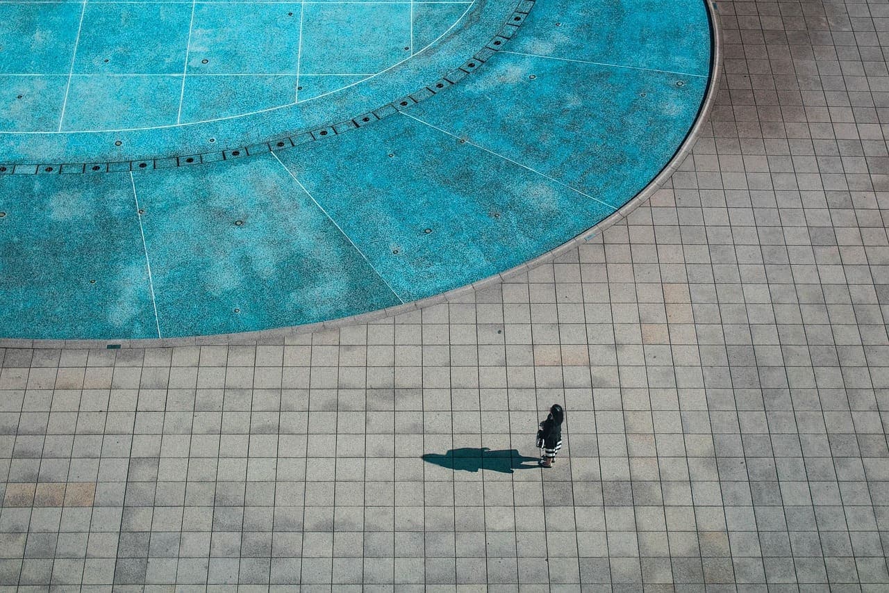 A round plunge pool seen from above, ringed by pale stone tiles, a lone bather crossing the deck