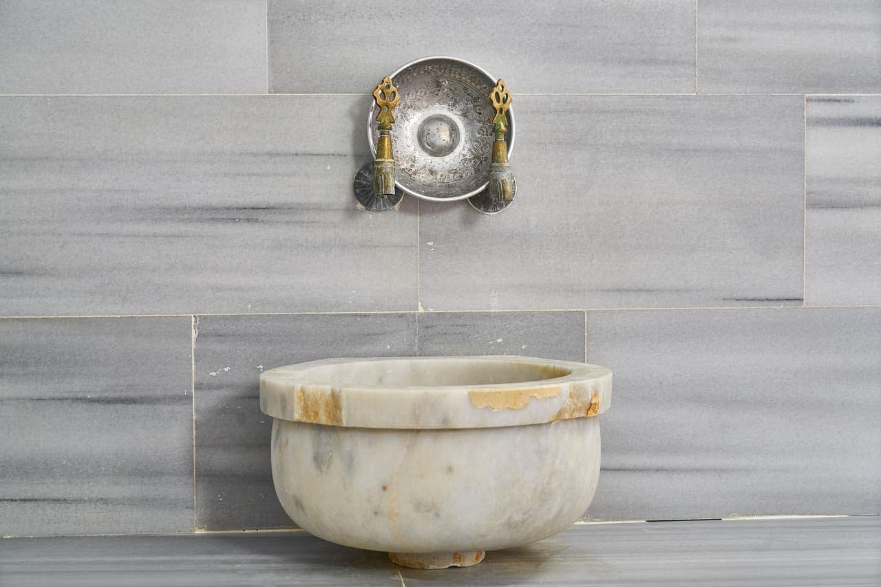 A carved marble basin under a brass tap on a grey marble wall